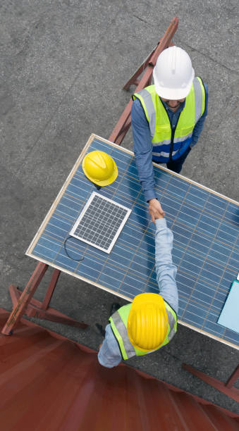 young engineer in hardhat and safety vest shake hands with project manager after agreeing a business plan. they use solar cell panel as a table. a large container is in the background. top view - renewable energy alternative energy technology solar energy imagens e fotografias de stock