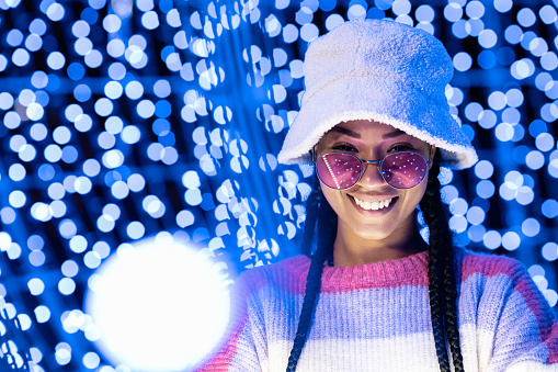 Joyful woman of color with a white beanie braided hairstyle and pink glasses enjoying the Christmas lights