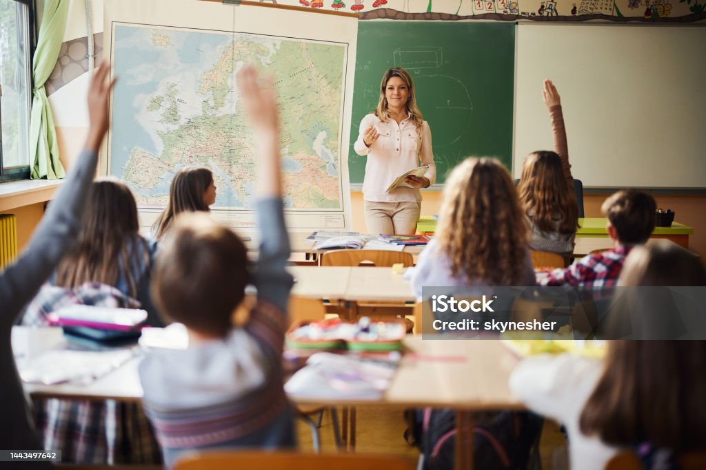 Who is going to answer my question? Smiling teacher aiming at her elementary students while wanting to hear the answers to her question. Teacher Stock Photo