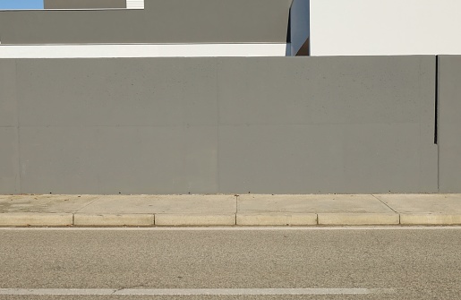 Gray concrete fencing wall with a modern house facade behind. Cement sidewalk and asphat road in front. Background for copy space.