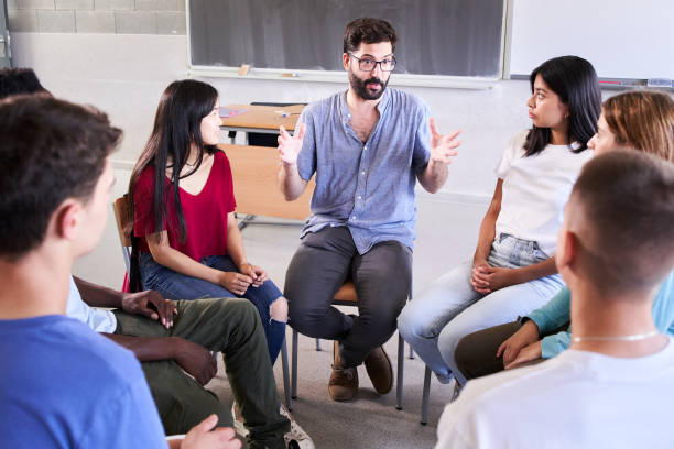 istock Diverse group of high school students sitting on chairs in a circle and interacting during a lesson, their Caucasian male teacher with them and talking. Education concept 1443500734