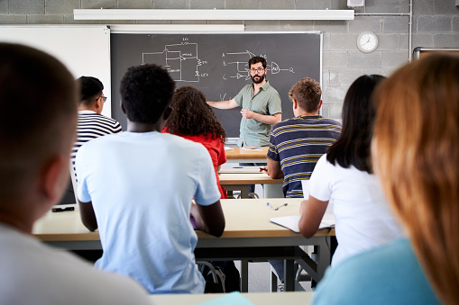 Male Tutor Teaching University Students In Classroom. Rear view from college attending at teacher in class. Professor explains the lesson on the blackboard. High quality photo
