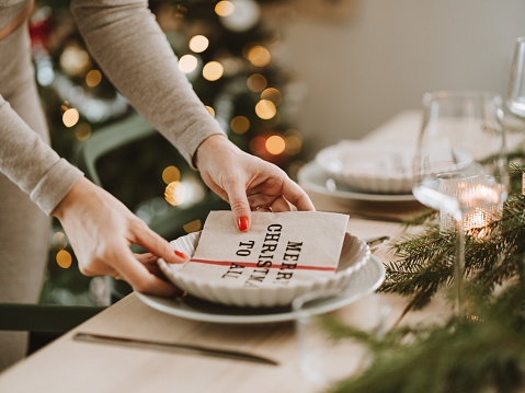 Woman setting the Christmas table preparing for dinner party.
Place setting on dining table with cutlery plate napkin and decorations for Christmas.