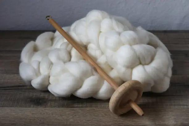 Wooden handspindle for spinning wool is laying on a skein of carded sheep wool on a woodden table, showcasing this traditional hobby of spinning.