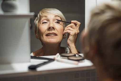 Mature woman is applying mascara in the bathroom.