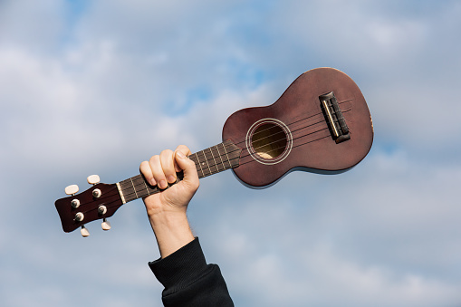 Guitar ukulele in man hand against cloudy sky, nature blurred background, copy space. Musical instrument for Folk music performed. Musical relaxation hobby concept.