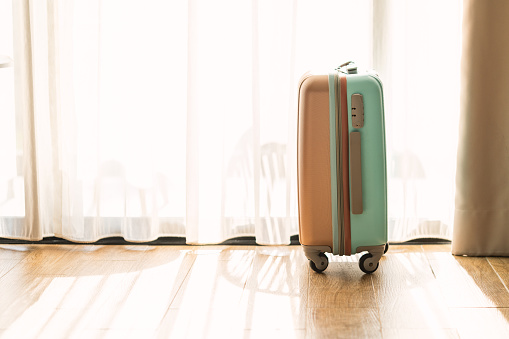 Side view small modern luggage with two tone color, stand on the wooden floor in a hotel room, background of white curtain, space for text and design.