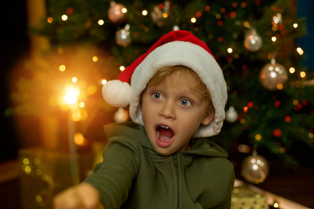 Cute boy in a New Year is hat, with big blue eyes and a mouth open in surprise holds a sparkling sparkler in his hand against the background of a decorated Christmas tree