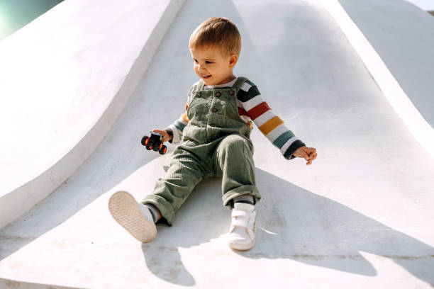 süßer kleiner junge, der mit einem autospielzeug in der hand auf einem spielplatz rutscht - child playground small toy stock-fotos und bilder