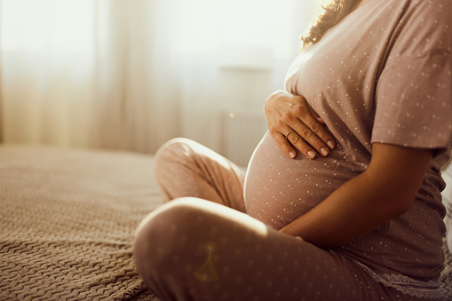 Close up of unrecognizable expecting woman holding her stomach while relaxing on a bed in the morning. Copy space.