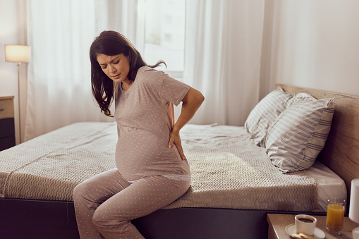 Sad pregnant woman holding her back in pain while sitting on a bed in the morning. Copy space.