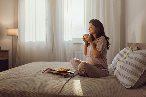 Smiling pregnant woman smelling fresh tea with her eyes closed while relaxing on a bed in the morning. Copy space.