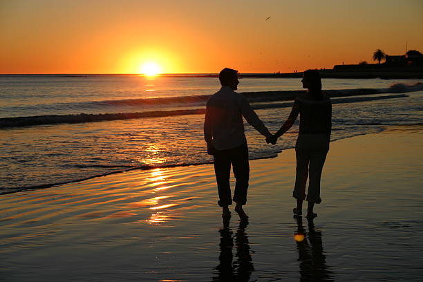 Loving couple walks hand-in-hand on the beach with a beautiful sunset in front of them
