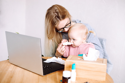 Portrait of young mother woman sitting at table near laptop, wooden box with paper napkins, holding little cute baby girl, trying to instill medicine into childs nose at home, toddler sniffing bottle.