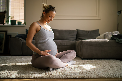 Beautiful young pregnant woman doing yoga and stretching at her home. She is woman with good and healthy habits and looks beautiful and happy.