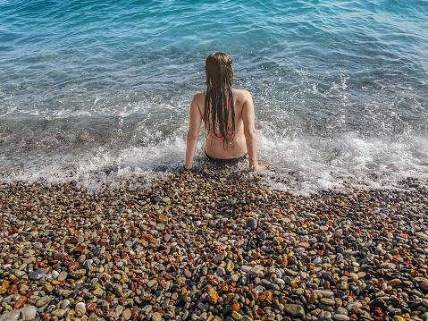 View from the back of a young woman in a swimwear and with wet long dark hair, sitting among the sea foam. Summer holidays on the shores of the Mediterranean Sea on a beach with multi-colored pebbles