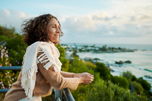 Smiling mature woman wearing a pashmina leaning on a railing and looking out at the ocean at sunset