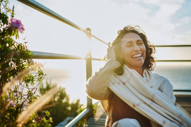 laughing mature woman sitting by the ocean at sunset - mulheres-maduras imagens e fotografias de stock