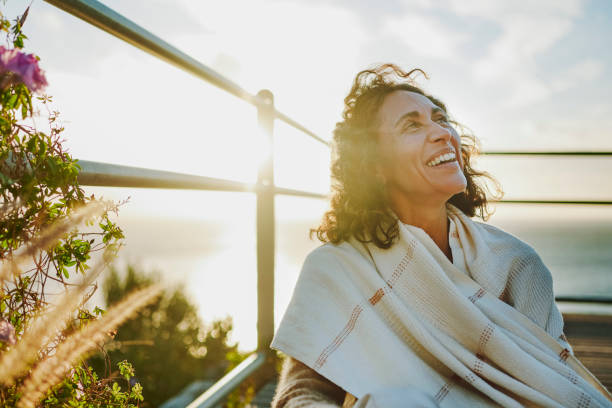 Mature woman laughing while sitting by the ocean at sunset