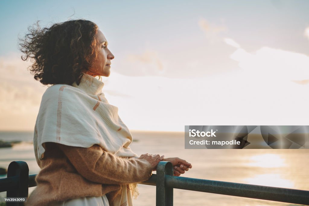 Mature woman watching the sunset over the ocean Mature woman wearing a pashmina leaning on a railing and looking out at the sunset over the ocean Contemplation Stock Photo