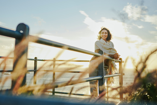 Portrait of a smiling mature woman wearing a pashmina leaning against a railing at a viewpoint by the ocean at sunset
