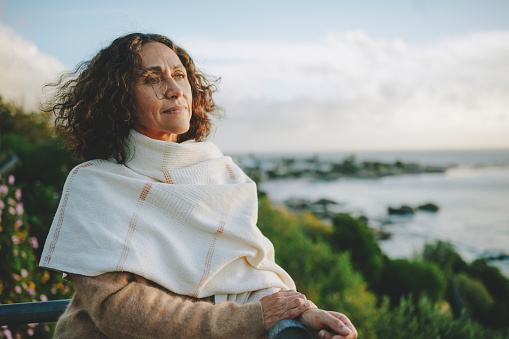 Mature woman wearing a pashmina leaning on a railing and looking out at the ocean at sunset