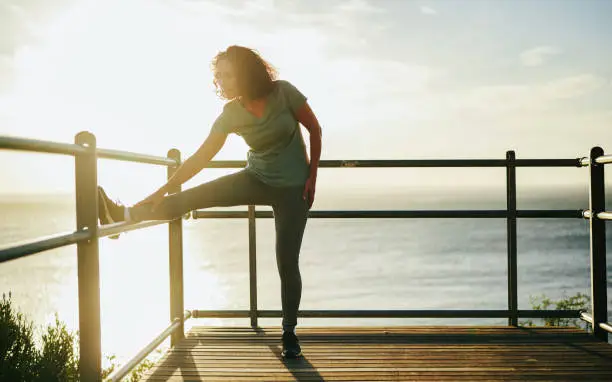 Fit mature woman in sportswear stretching on the railing of a scenic viewpoint before a run by the ocean at sunset