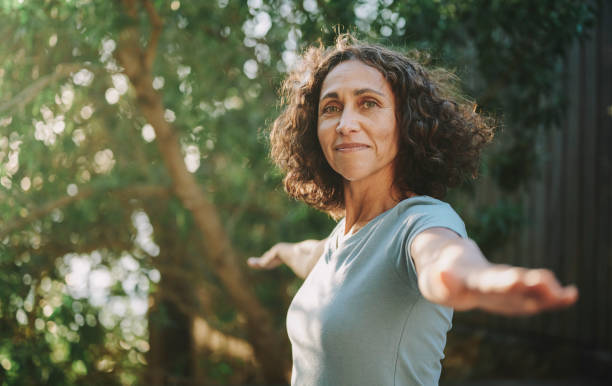 Smiling mature woman doing yoga outside in a park in summer Portrait of a smiling mature woman practicing yoga outside in a park in the summertime warrior position stock pictures, royalty-free photos & images