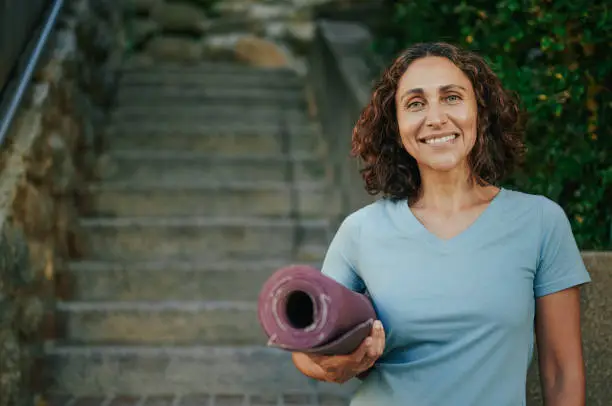 Portrait of a smiling mature woman in sportswear standing with an exercise mat before yoga in a park in summer