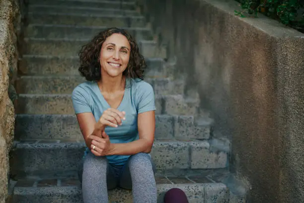 Portrait of a smiling mature woman in sportswear sitting on stairs with her exercise mat outside before yoga in a park in summer