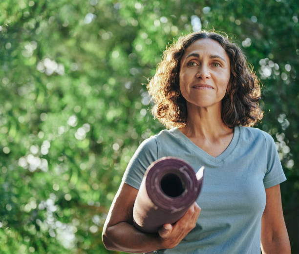 mujer madura sonriente cargando una colchoneta de ejercicios de pie en un parque - colchoneta de ejercicio fotografías e imágenes de stock