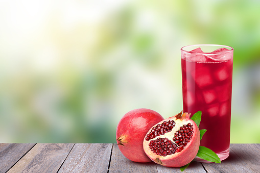 Glass of pomegranate juice with fresh fruit on wooden table with green nature blurred background. Copy space for text.