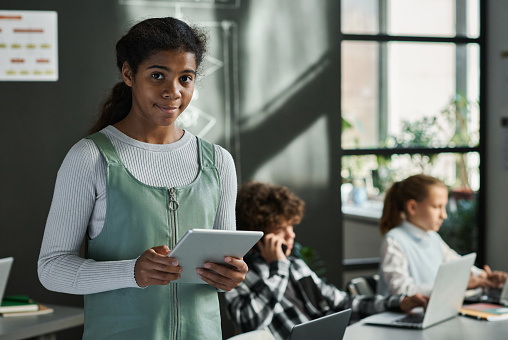 Portrait of African American schoolgirl with tablet pc looking at camera standing at classroom with her classmates in background
