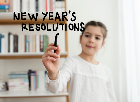 cheerful little girl writing new year's resolutions with a black pen on the transparent wipe board at home