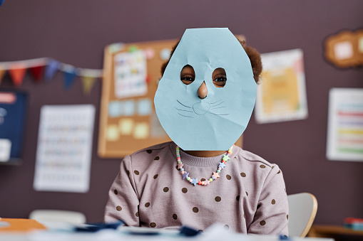 Cute little girl with blue paper mask on face sitting in front of camera at lesson in classroom of nursery school and looking at you