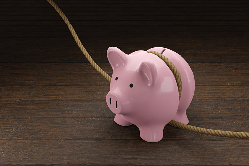 Pink piggy bank is tightened by a rope on wooden table. Illustration of the concept of budget tightening and reduction of spending