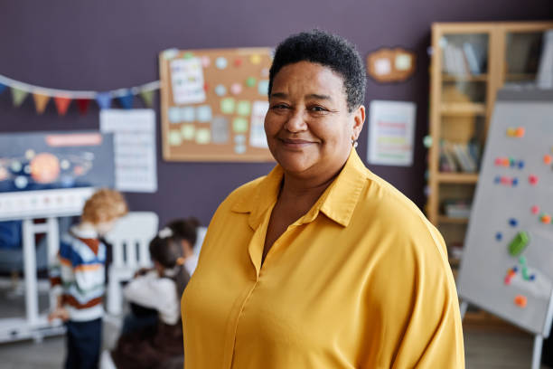 Successful professional teacher of nursery school looking at camera Successful professional mature female teacher of nursery school looking at camera while standing against group of little learners instructor stock pictures, royalty-free photos & images