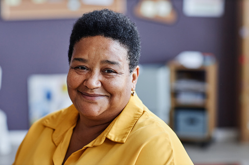 Mature smiling black woman in yellow blouse sitting in front of camera and looking at you against school supplies in classroom