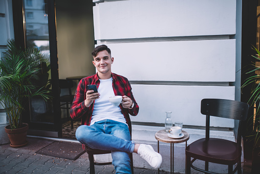 Positive young man wearing casual clothes smiling and sitting with crossed legs on chair in street cafe while drinking coffee and browsing smartphone while looking at camera