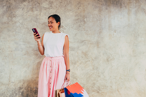 A smiling Asian female holding shopping bags and reading something on her mobile phone after buying some things at the store. She is standing against white background.