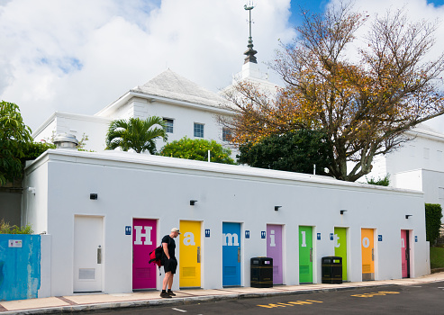 Hamilton, Bermuda-May 24, 2022- A tourist waits for his companion in front of  a row of colorful rest room doors that spell out the capitol city's name that are located near Hamilton City Hall and Victoria Gardens.