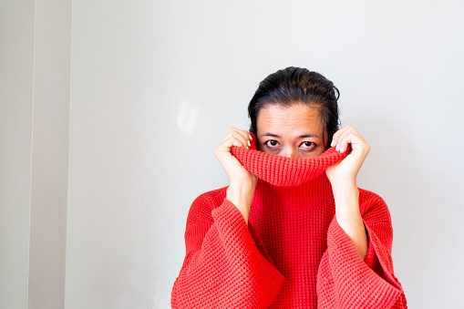 Woman in a red sweater pulls the turtleneck up over her head looking at the camera, symbolizing hiding and protecting herself  from the world