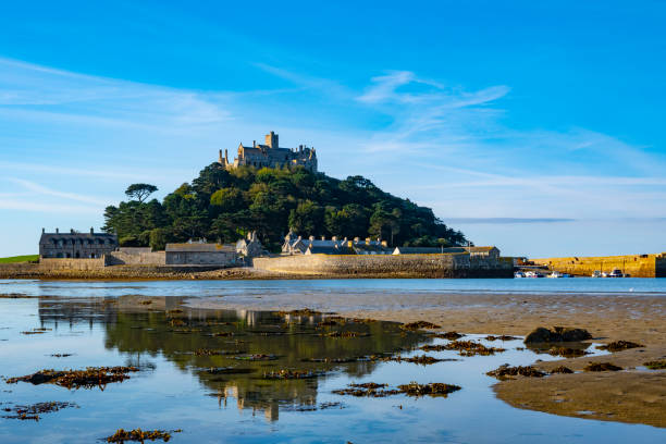 St Michaels Mount Castle. Close up picture of old stone castle with lake in the background. The sky above the castle is gray and overcast. The remnants of a rock wall are in the foreground of the picture. There is moss growing on the rocks in the foreground and on the castle. The overall color tone is very gray.