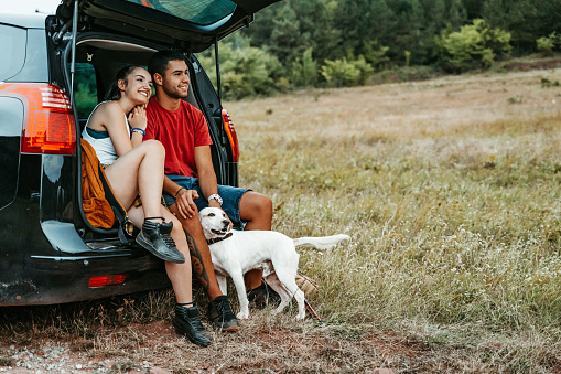 Beautiful young hiking couple petting their dog while sitting in a car trunk.