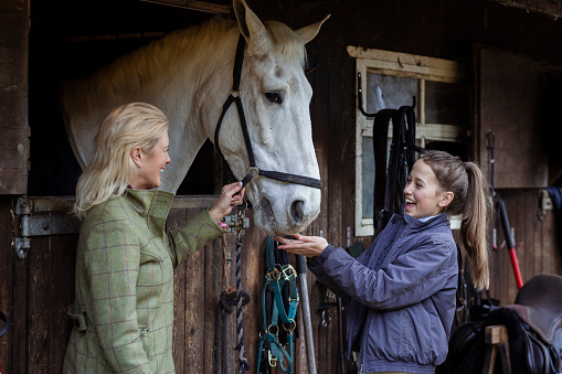 A woman and her daughter standing outdoors at their stable at home in Newcastle Upon Tyne, England. They are petting and feeding their pet horse while they bond with each other.