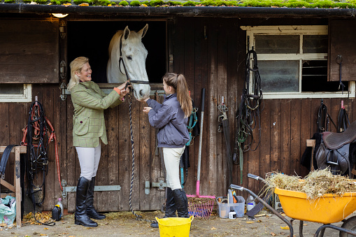 A woman and her daughter standing outdoors at their stable at home in Newcastle Upon Tyne, England. They are petting and feeding their pet horse while they bond with each other.