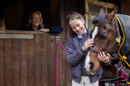 A female teenager standing outdoors at her stables at home in Newcastle Upon Tyne, England. She is looking at her horse and smiling while stroking it. Her mother is smiling while watching her from the stables.