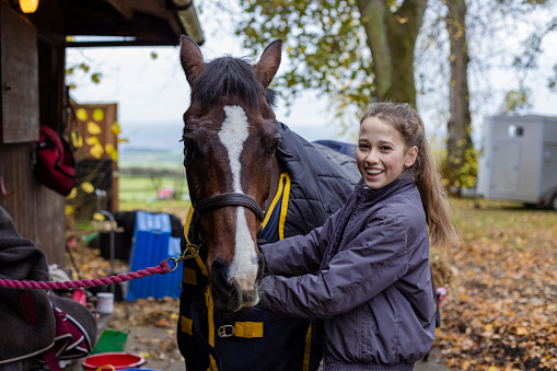 A female teenager standing outdoors at her stables at home in Newcastle Upon Tyne, England. She is looking at the camera and smiling while caring for her horse, who is fixed to the stable with a rope.