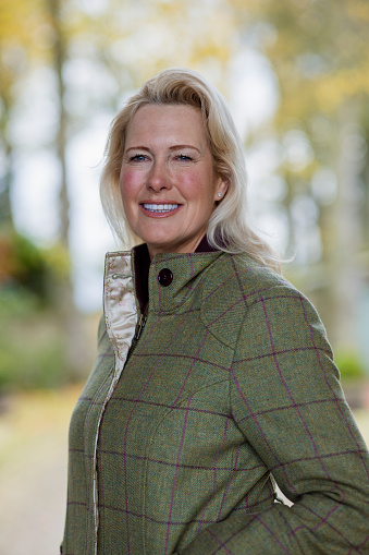 A portrait of a mature woman standing outdoors in a rural scene in Newcastle upon Tyne, England, wearing a tweed jacket while looking at the camera and smiling.