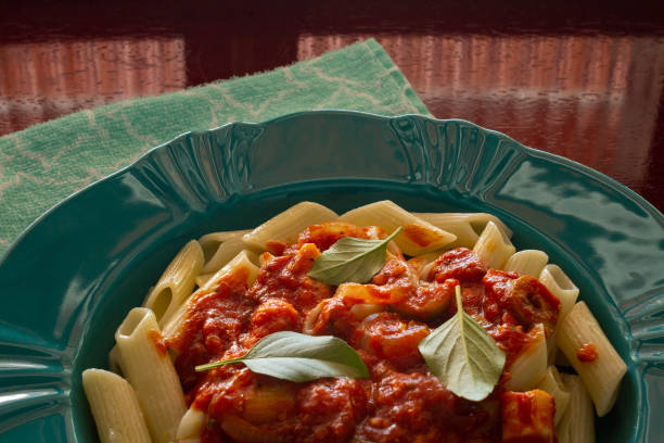 Stock photo showing close-up, elevated view of a red plate containing a meal of freshly cooked penne pasta tossed in a homemade marinara sauce made with tomatoes, garlic, olive oil, and herbs. The meal is topped with red cherry tomatoes, torn pieces of buffalo mozzarella cheese and basil leaves.